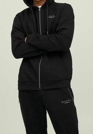 Sudadera negra con cremallera y pantalones de chándal a juego, confeccionados en un tejido suave, con capucha ajustable mediante cordón y detalle de logo bordado en el pecho y el muslo.