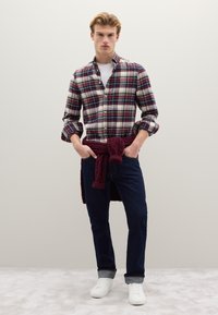 Camisa de cuadros en azul marino, rojo y blanco; vaqueros oscuros; zapatillas blancas; suéter tejido burdeos atado a la cintura; mangas largas remangadas.