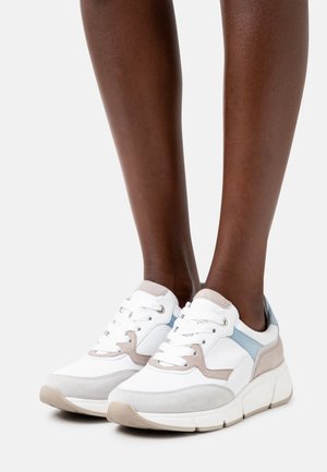 Jambes portant des baskets blanches avec des accents beige et bleu clair, semelles épaisses et lacets blancs sur un fond blanc.