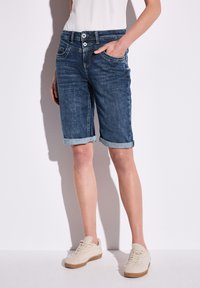 Bermudas de mezclilla en lavado oscuro con puños doblados, cuentan con dos bolsillos frontales y una cremallera. Combinadas con zapatillas de textura clara.