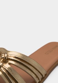 Sandale plate métallique dorée avec plusieurs fines lanières nouées ensemble sur une semelle beige, sur fond blanc.