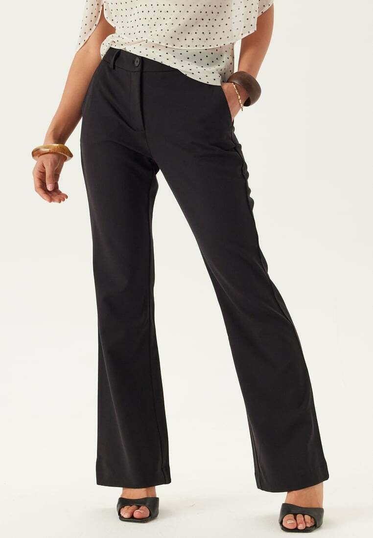 Vrouw die zwarte rechte broek draagt, witte blouse met zwarte stippen en zwarte open-toe hakken, staand met één hand in de zak.