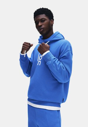 Jeune homme ajustant les cordons blancs d'un sweat à capuche bleu porté avec un pantalon bleu assorti et une chemise blanche en dessous, sur un fond uni.
