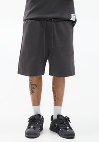 Grijze shorts met zijzakken, een verstelbare tailleband en zichtbare stiksels. Draagt zwarte sneakers en witte crew-sokken.