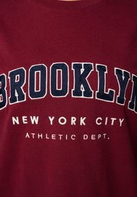 Bordeaux sweatshirt met witte en marineblauwe geborduurde tekst: "BROOKLYN NEW YORK CITY ATHLETIC DEPT." Ronde halslijn, katoenen stof.