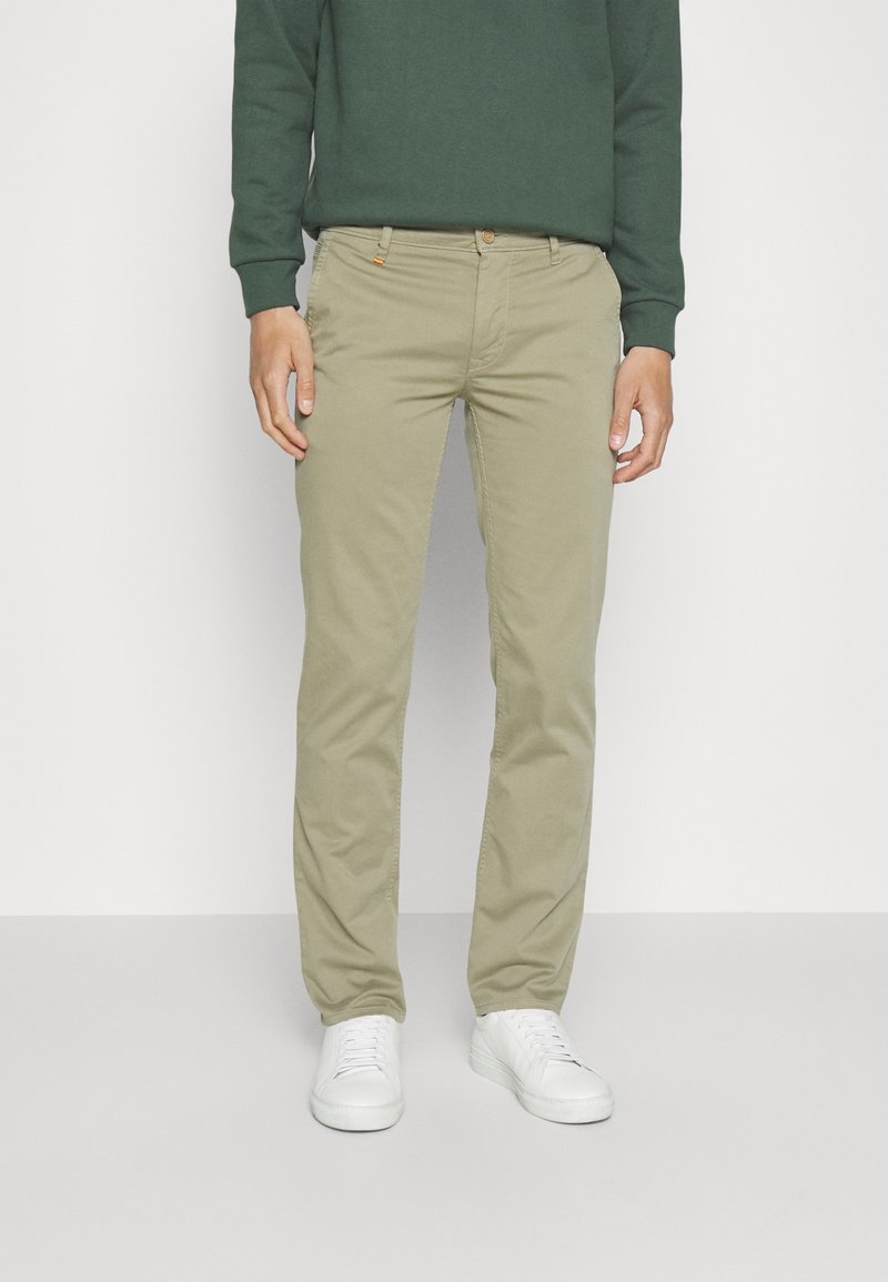 Beige chinos slim fit, joissa on nappikiinnitys ja etutaskut, yhdistettynä tummanvihreään collegepaitaan ja valkoisiin lenkkarikenkiin.