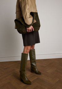 Stivali al polpaccio in pelle verde oliva con punta a punta e tacchi a blocco, abbinati a una gonna marrone plissettata e a una borsa a spalla in suede verde scuro.