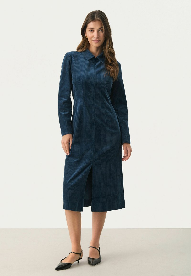 Langärmliges, marineblaues Cordkleid mit Kragen, frontalem Schlitz und geradem Schnitt. Kombiniert mit schwarzen Slingback-Schuhen mit Absatz.