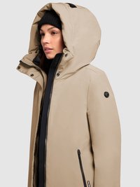 Beige isoleret jakke med stor hætte, sorte detaljer og markante lynlåse. Har sidelommer og en glat tekstur.