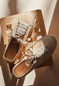 Zapatos de ante beige con cordones de tela, punta redonda, bordes cosidos y suela tejida, descansando sobre una superficie de madera con una iluminación suave.