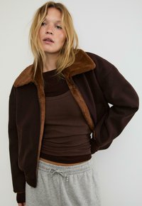 Brahna zip-up jakna z umetnim krznom na ovratniku, položena čez rjav top brez rokavov, kombinirana s sivo trenirko. Teksturirana tkanina, sproščen kroj.
