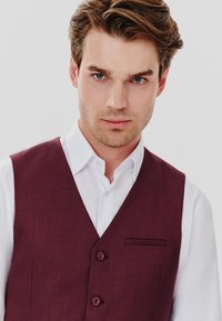 Bordowy kamizelka wykonana z teksturowanej tkaniny z dwiema guzikami, z małą kieszonką, noszona na białej koszuli.