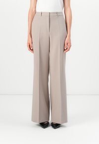 Pantaloni a gamba larga in tessuto beige chiaro con una texture liscia, dotati di una piega frontale e vita aderente. Abbinati a scarpe nere con punta appuntita.