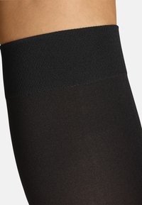 Collant neri opachi con una fascia elastica testurizzata in vita. Il materiale è liscio, passando da una parte superiore più spessa a una sezione gamba più sottile.
