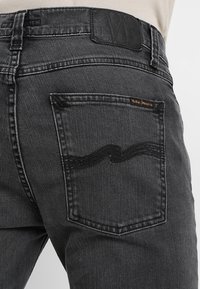 Černé denimové džíny s texturovanou zadní kapsou a vlnitým švem, s koženou nášivkou na pasu. Klasický pět-kapsový styl.