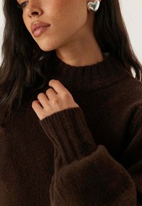 Maglione marrone lavorato a maglia con un collo alto e polsini a coste, caratterizzato da una texture morbida. Il modello indossa orecchini a forma di cuore in argento.