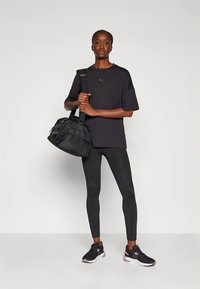 Svart atletisk t-skjorte, løstsittende, kombinert med svarte leggings og svarte joggesko med rosa detaljer. Modellen holder en liten svart bag.