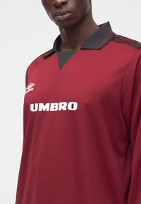 Άνδρας που φορά σπορ μπλούζα Umbro μπορντό με μαύρο γιακά και διακοσμητικό μοτίβο στον ώμο, εμφανιζόμενος από το στήθος μέχρι το πηγούνι.