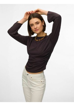 Kvinde med kort brunt hår iført en mørk langærmet top, hvide bukser og en perlehalskæde, poserer med armene løftet bag hovedet.