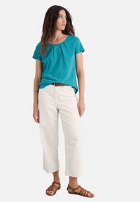 Tyrkysový top s krátkými ruk ávy a nařasením, spárovaný s bílými širokými kalhotami a hnědými sandály na pásku. Hladký materiál s mírným leskem.