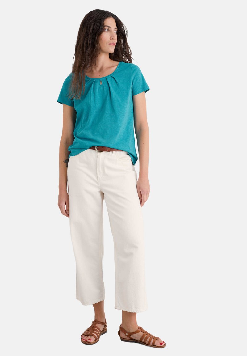 Tyrkysový top s krátkými rukávy a nařasením, spárovaný s bílými širokými kalhotami a hnědými sandály na pásku. Hladký materiál s mírným leskem.