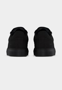 Ζευγάρι μαύρα χαμηλά αθλητικά παπούτσια, εμφανιζόμενα από την πίσω πλευρά, με παχιά ελαστικά πέλματα και επενδεδυμένα κολάρα, σε λευκό φόντο.