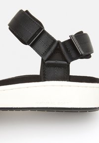 Svart sandal med texturerade nylonremmar, en vadderad svart fotbädd och en vit gummisula prydd med "NATURAL SOFT™."