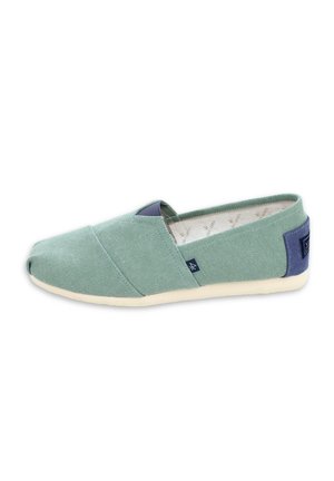 Zapato tipo slip-on de tela canvas verde claro con suela beige y detalle de talón de tela azul oscuro, visto de lado sobre fondo blanco.