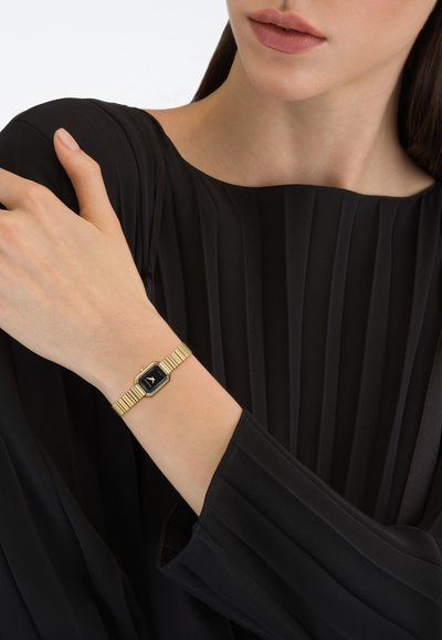 Aranyszínű karóra hatszögletű számlappal és fekete háttérrel. Texturált fém karkötővel és redős fekete blúzzal.