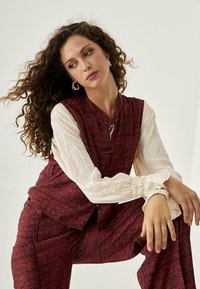 Top sin mangas de color marrón con patrón, con un cuello alto, usado sobre una blusa de textura crema. El conjunto incluye pantalones holgados con detalles de bolsillos.