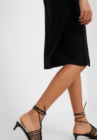 Robe noire à mi-mollet avec une fente sur le côté, associée à des sandales noires à talons hauts et à lanières, mettant en valeur une texture lisse et un design épuré.