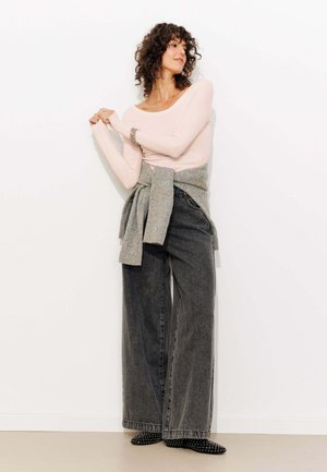 Haut rose clair à manches longues, pull gris noué à la taille, jeans larges gris foncé et escarpins noirs à clous. Fond blanc uni.