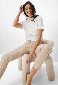Witte T-shirt met korte mouwen en beige aansluitende broek, zittend op een zachte crème kleurige stoel. Het model draagt een geprinte sjaal en zilveren sieraden.
