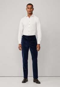 Bijela košulja s dugim rukavima i smeškastim dugmadima uparena s tamnoplavim kordura hlačama, nosi se s tamnosmeškim cipelama.