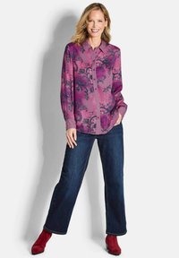 Langärmlige rosa Bluse mit schwarz floralem Muster, Knopfleiste vorne und strukturierten Akzenten. Kombiniert mit dunklen, weit geschnittenen Jeans und roten Knöchelstiefeln.