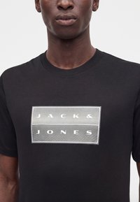 Άνδρας που φοράει μαύρο t-shirt με λευκό ορθογώνιο λογότυπο που εμφανίζει το "JACK & JONES" με κεφαλαία γράμματα.