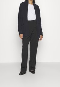 Marinblå stickad kofta med öppen framsida, ribbad textur och sidoficka, i kombination med vit t-shirt och svarta skräddarsydda byxor.