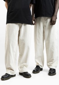 Wide-legged, off-white corduroy pants with subtle texture, paired with black shoes featuring yellow stitching, worn by two individuals.