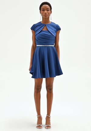 Donna che indossa un vestito blu a maniche corte, lungo fino al ginocchio, con una cintura argento e sandali argentati con tacco alto, in piedi davanti a uno sfondo semplice.