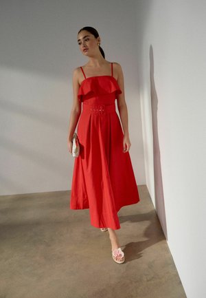 Kobieta spacerująca wewnątrz pomieszczenia, ubrana w czerwoną sukienkę midi z paskiem i falbanką na górze, trzymająca beżową kopertówkę.