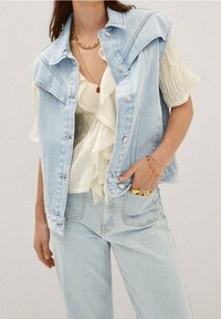 Helles blaues Denim-Westen mit strukturiertem Kragen, Edelstahlknöpfen, darunter eine mehrlagige weiße Rüschbluse und passende helle Jeans.
