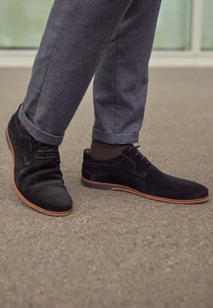 Zapatos de ante negro con un diseño elegante, suelas marrones contrastantes y detalles sutiles de cordones, combinados con pantalones grises, sobre una superficie texturizada.