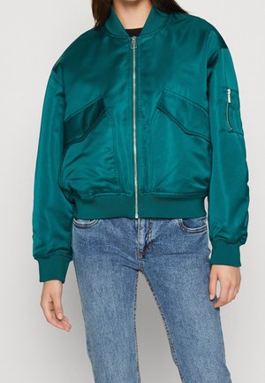 Personne portant une veste bomber en satin bleu sarcelle avec fermeture éclair à l'avant et poches à rabat inclinées, associée à un jean en denim bleu clair.