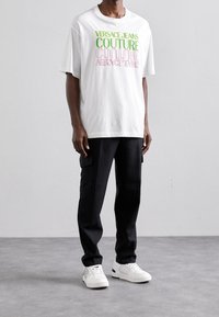 Bílý bavlněný tričko s multicolor nápisem "Versace Jeans Couture", spárované s černými cargo kalhotami a bílými teniskami.