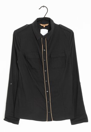 Sort langærmet skjorte med knapper og brystlommer samt beige piping langs knaplinjen, hængende på en sort bøjle.