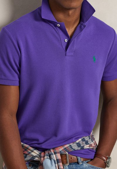 Μωβ μπλούζα πόλο με υφή υφάσματος, δύο κουμπιά στον γιακά και ένα μικρό πράσινο λογότυπο. Ένα καρό πουκάμισο είναι δεμένο γύρω από τη μέση.