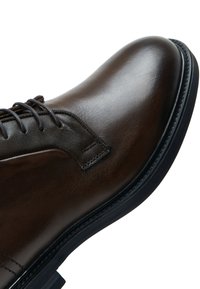 Primer plano de un zapato de vestir de cuero marrón oscuro pulido, con cordones negros y suela negra, sobre fondo blanco.
