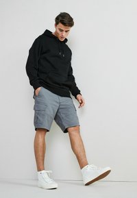 Fekete kapucnis hoodie zsinórokkal, szürke cargo shortszabak oldalsó zsebekkel, és fehér magasszárú sportcipők textúrált talppal. Kényelmes öltözet bemutatva.