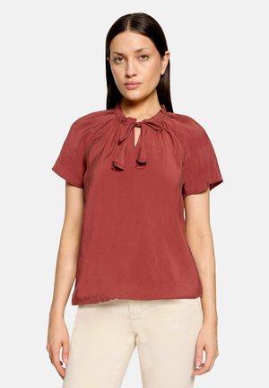 Vrouw met steil donker haar draagt een roestrode kortermige blouse met een gestrikte halslijn en crème gekleurde broek, staand tegen een effen achtergrond.