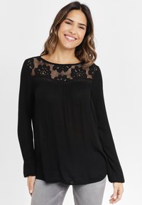 Svart långärmad blus med spetsvid yoke som har blommönster. Mjuk textur, avslappnad passform och rynkor fram. Matchas med grå jeans.